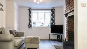 A living room with a grey sofa, a fluffy ottoman, a flat-screen TV on a stand, a fireplace, and a window with patterned curtains. - Henllys (Harlech)