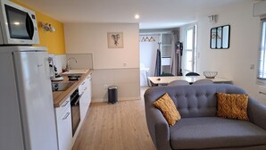 A compact living space with a kitchenette, a grey sofa with patterned cushions, and a dining area with a table and chairs. - Appartement Refait à Neuf Tout Équipé en RDC Avec Jardin Terrasse ,barbecue (Avrillé)
