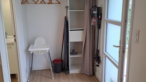A compact room with a white chair, a small table, a broom, a dustpan, and a trash bin. There is a window with a curtain, a door, and a coat rack with hangers. - Appartement Refait à Neuf Tout Équipé en RDC Avec Jardin Terrasse ,barbecue (Avrillé)