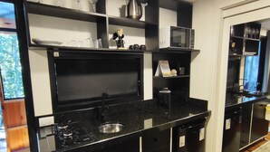 A modern kitchen with black cabinetry, a stainless steel sink, and a microwave. There are shelves above the counter holding glasses and a kettle. - Cabanas Loft Paraíso (Gramado)