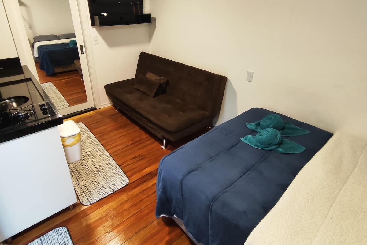 A compact room with a kitchenette, a sofa, a bed, and a flat-screen TV mounted on the wall.