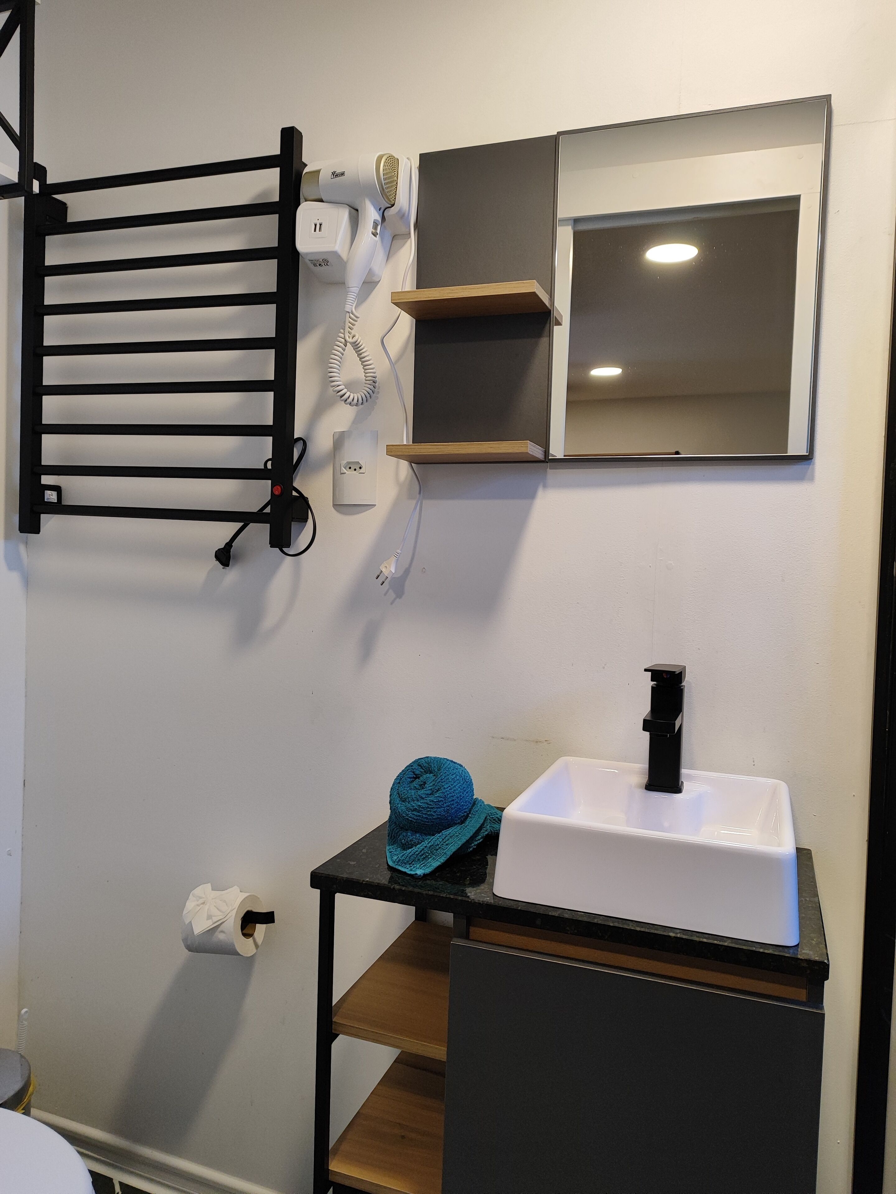 Un baño con toallero negro, lavabo blanco con grifo negro, espejo y toalla en un estante.