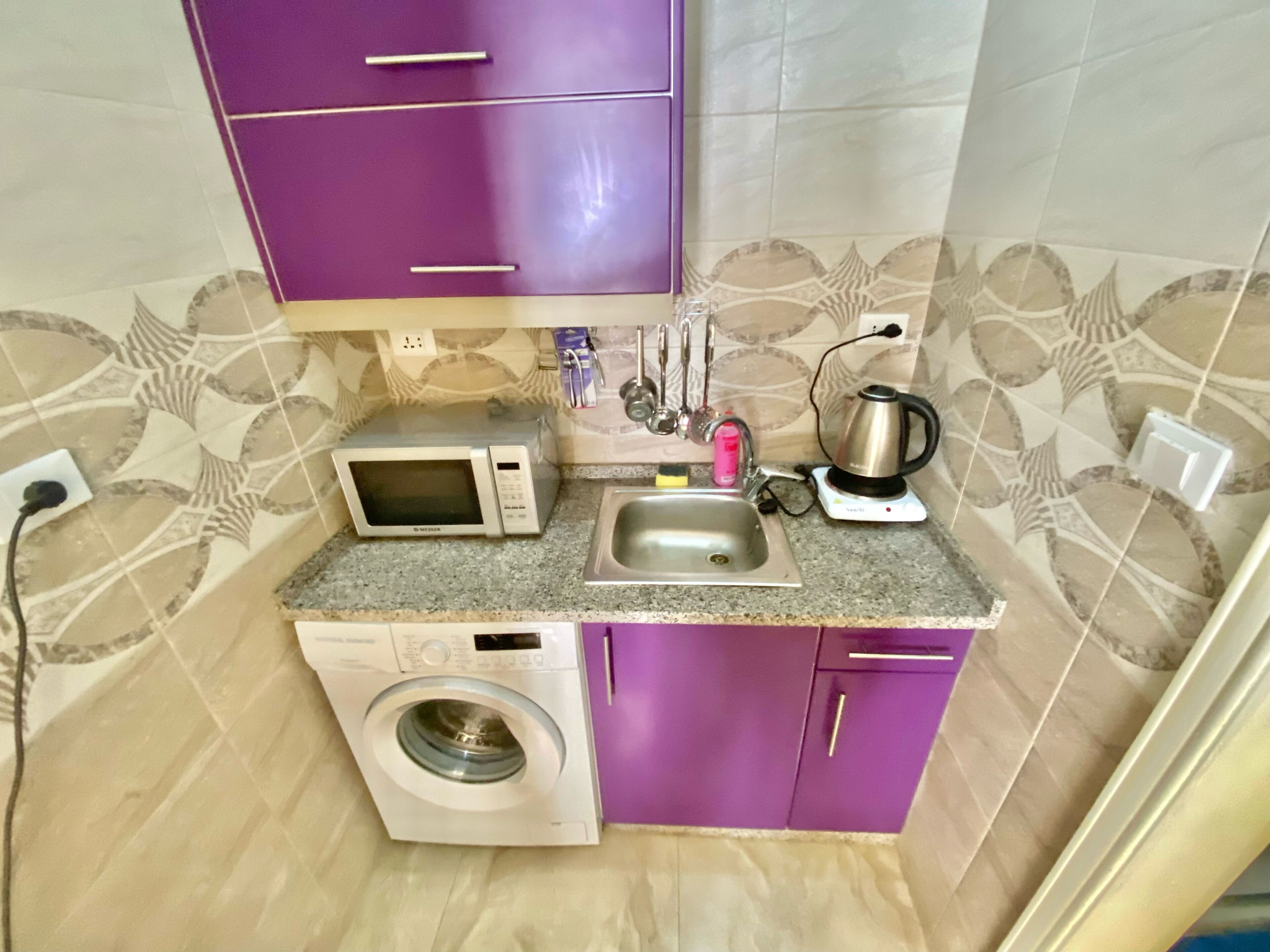 A compact kitchen with a washing machine, a microwave, a kettle, and a sink.