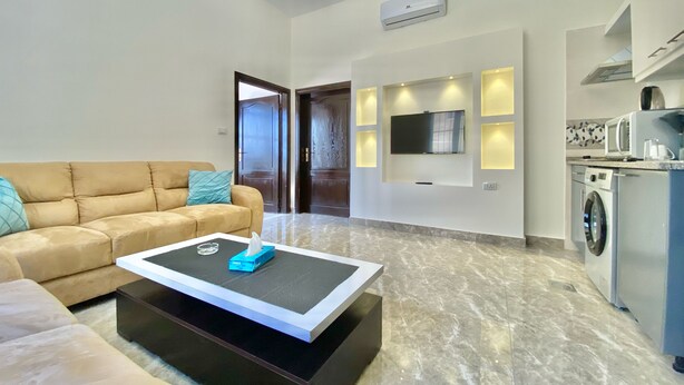 A modern living room with a sofa, a coffee table, a TV mounted on the wall, and a kitchen area with appliances.