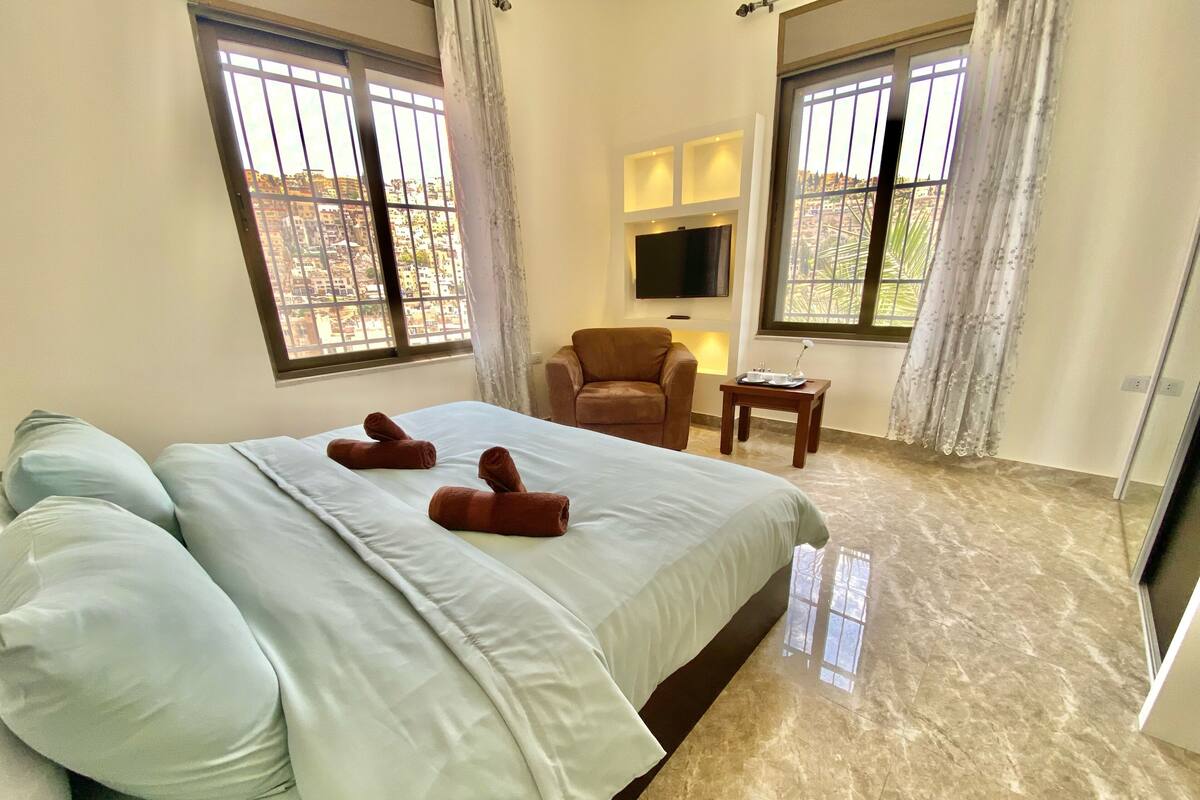 A hotel room with a large bed, two windows with curtains, a television, a small table with a lamp, and a chair.