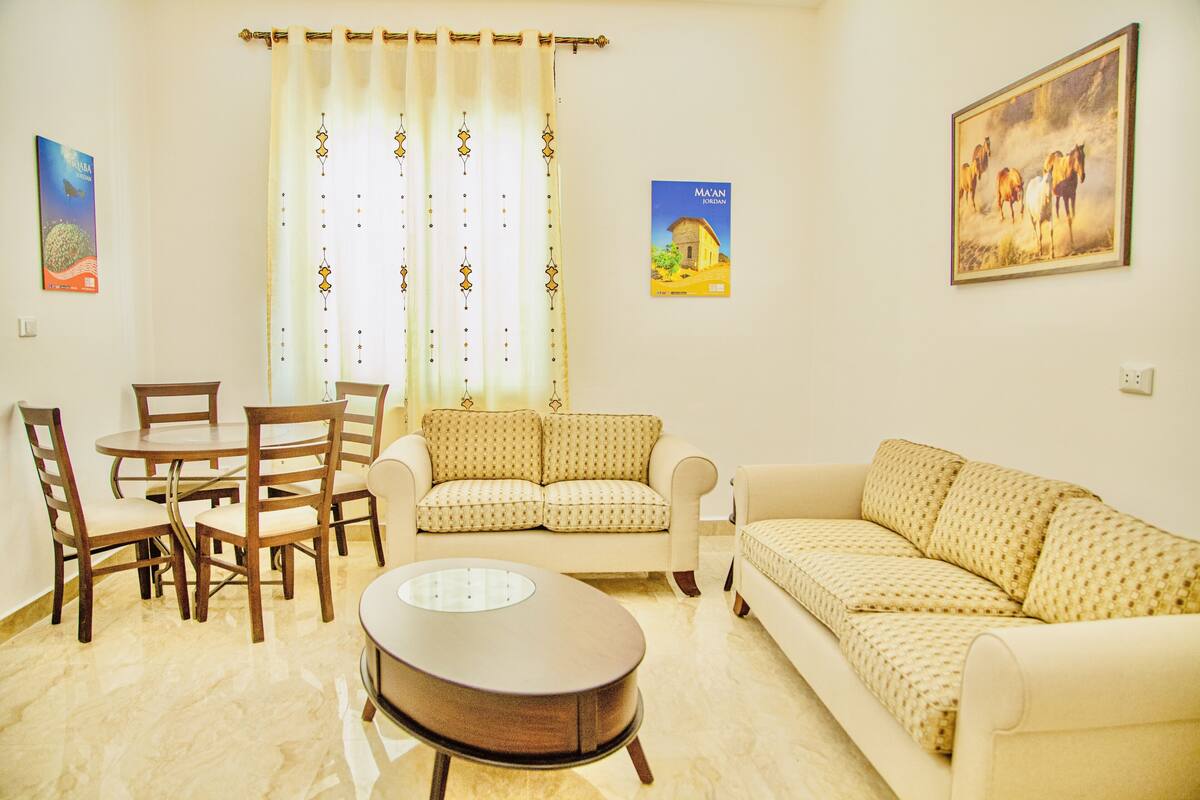 A living room with a sofa, two armchairs, a coffee table, and a dining table with chairs. There are curtains on the window, a painting on the wall, and a poster on the wall.