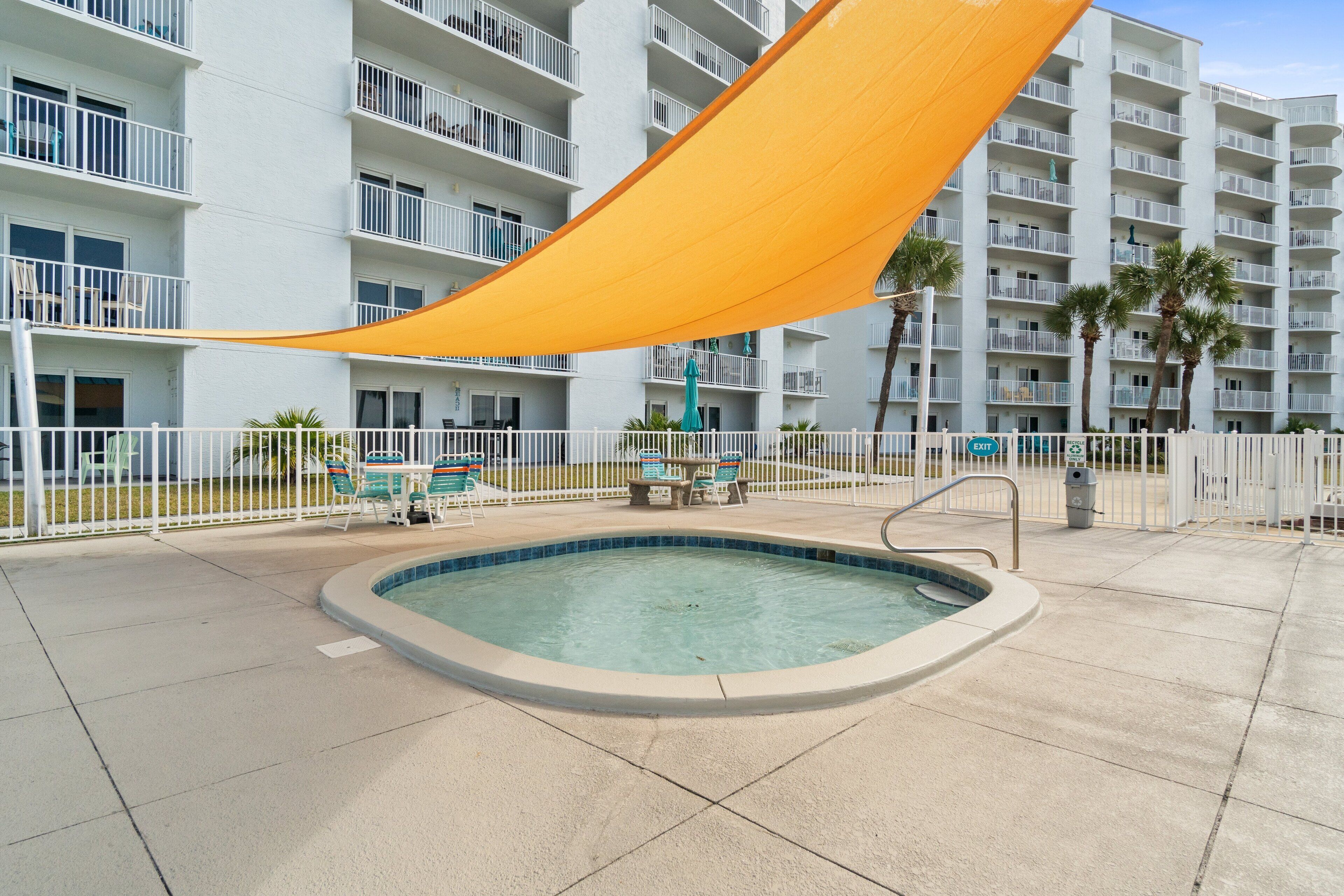 A swimming pool with a sunshade, surrounded by a white fence, and adjacent to a multi-story building with balconies.