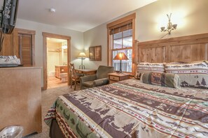 A bedroom with a large bed, wooden headboard, a desk with a chair, and a nightstand with a lamp.