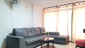 Ein Wohnzimmer mit einem grauen Sofa, einem kleinen runden Tisch mit einer Vase und einem Wandventilator.