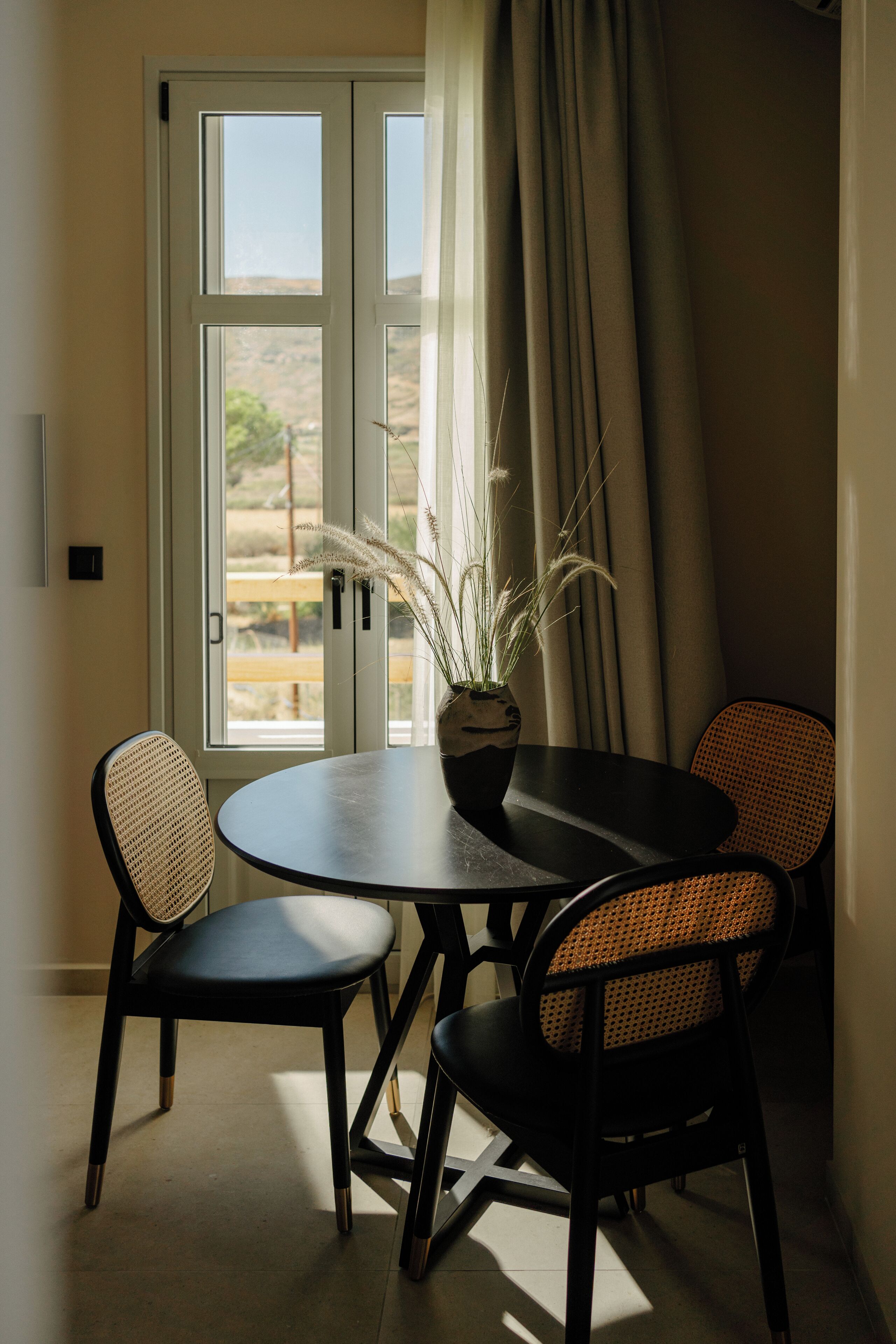Un comedor con una mesa redonda y dos sillas, una planta en maceta y vistas a través de una ventana con cortinas.