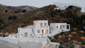 A modern white villa with a unique architectural design, featuring multiple levels and a spacious outdoor area with a view of the surrounding hills. - Pnoes Skyros (Skiros)