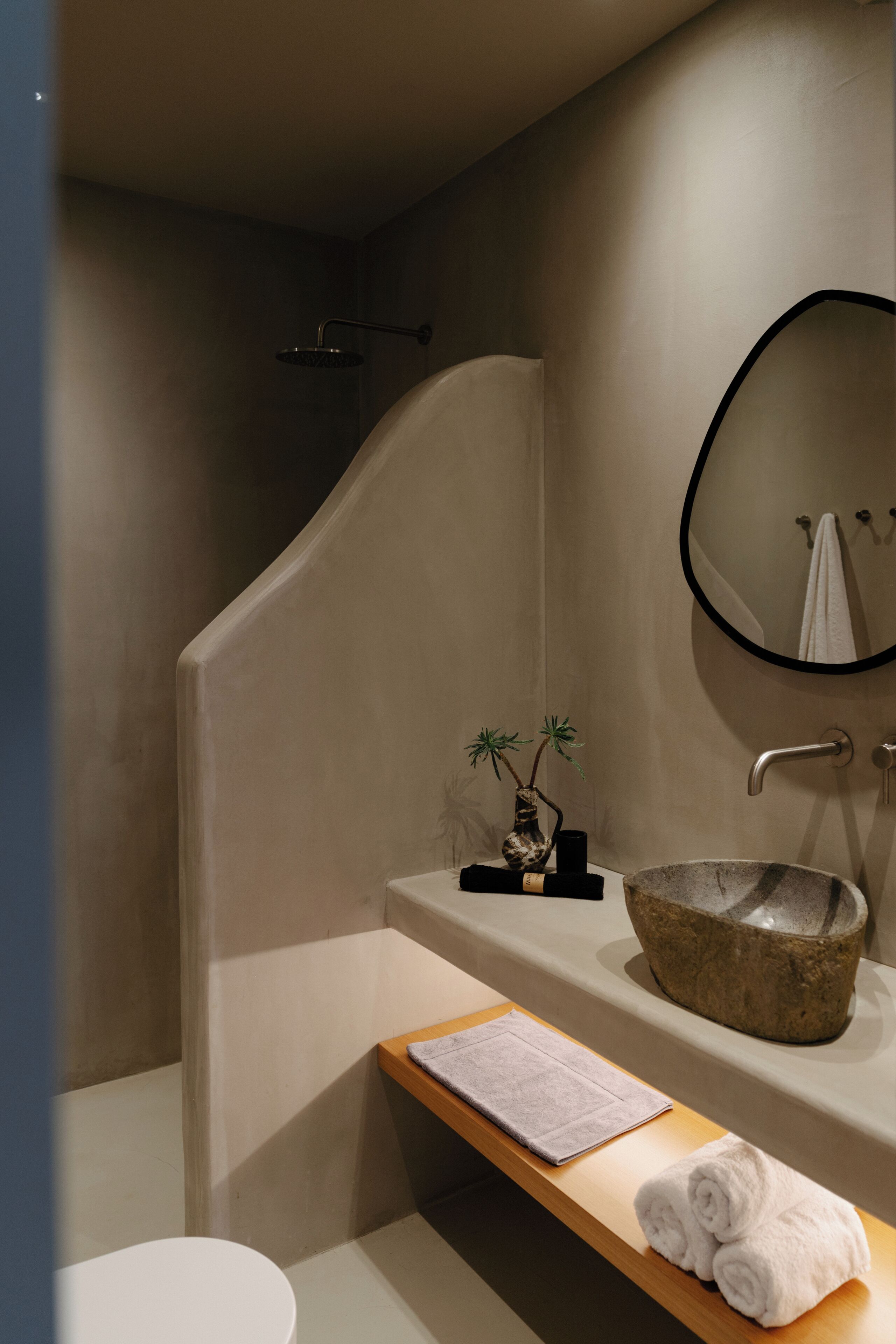 Un baño moderno con una bañera independiente, un espejo redondo y un estante de madera con toallas y un pequeño florero.