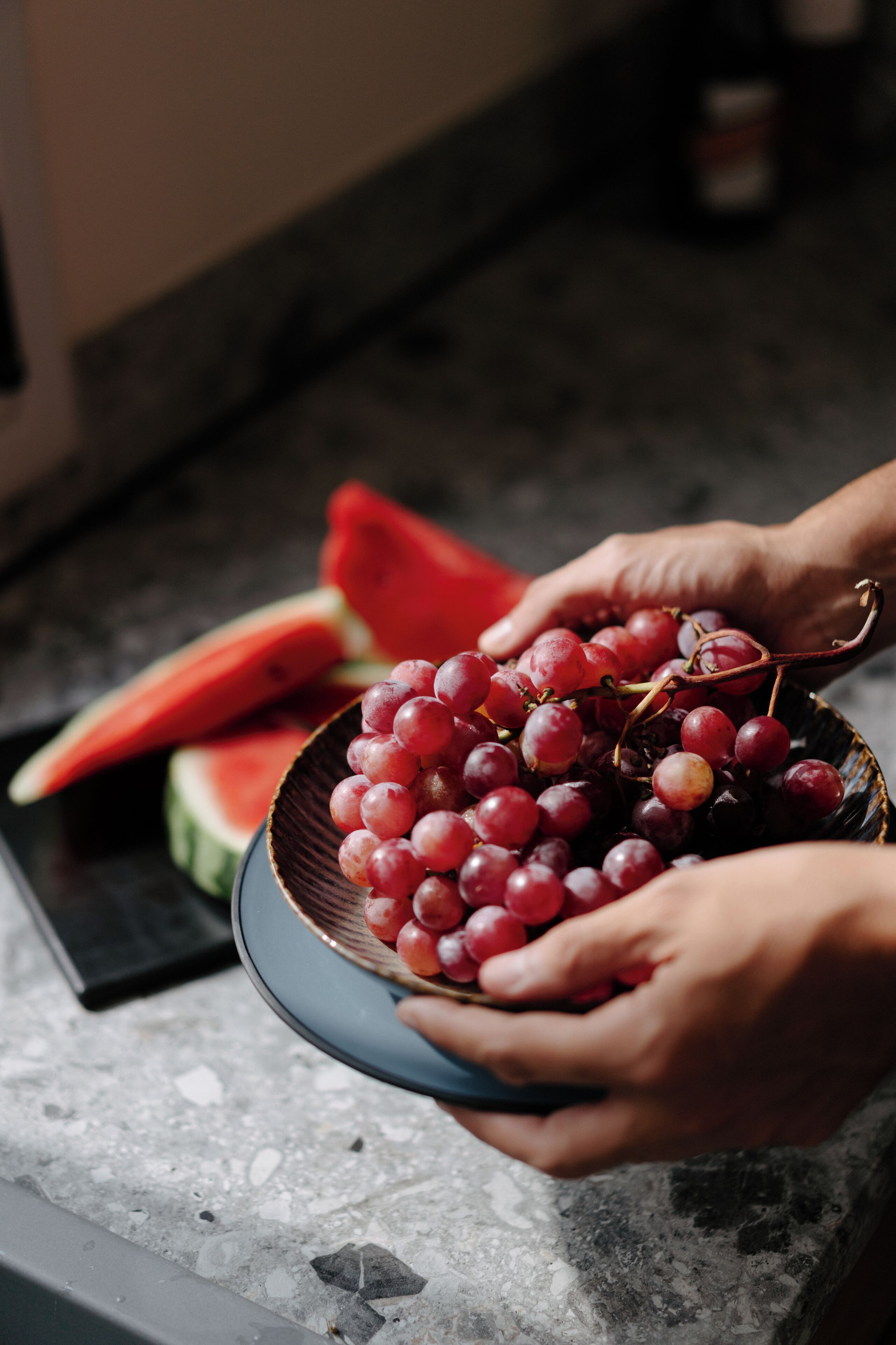 A person holding a bowl of red grapes and a slice of watermelon on a kitchen counter.