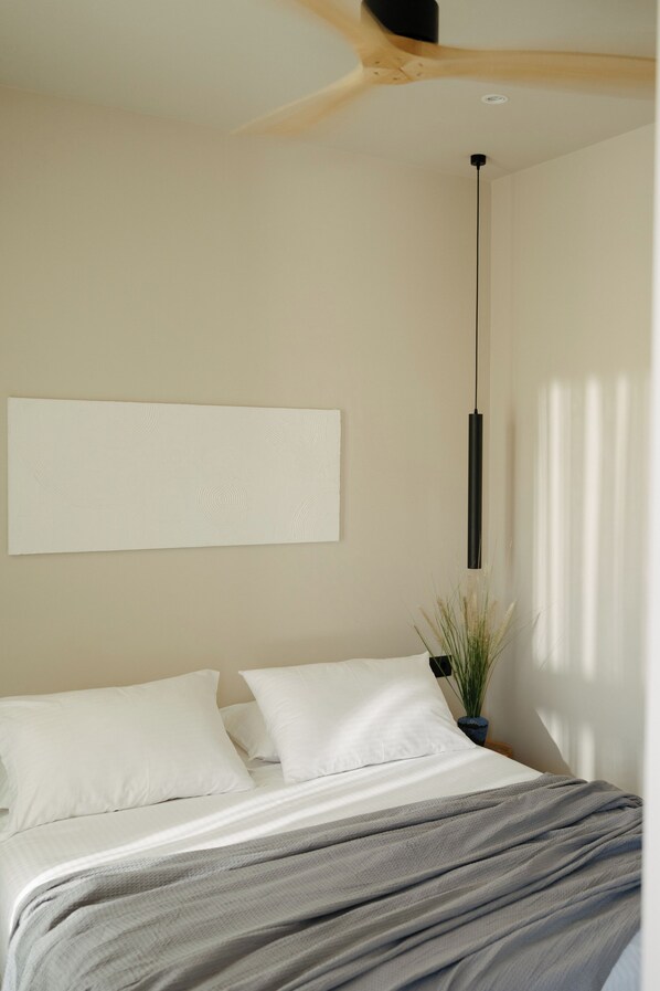 Un dormitorio con una cama, almohadas blancas, una manta gris, una lámpara colgante y una planta en maceta.