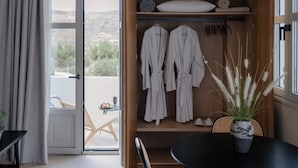 A room with a wooden wardrobe containing white robes, a small table with a potted plant, and a view of a balcony with a chair and table. - Pnoes Skyros (Skiros)