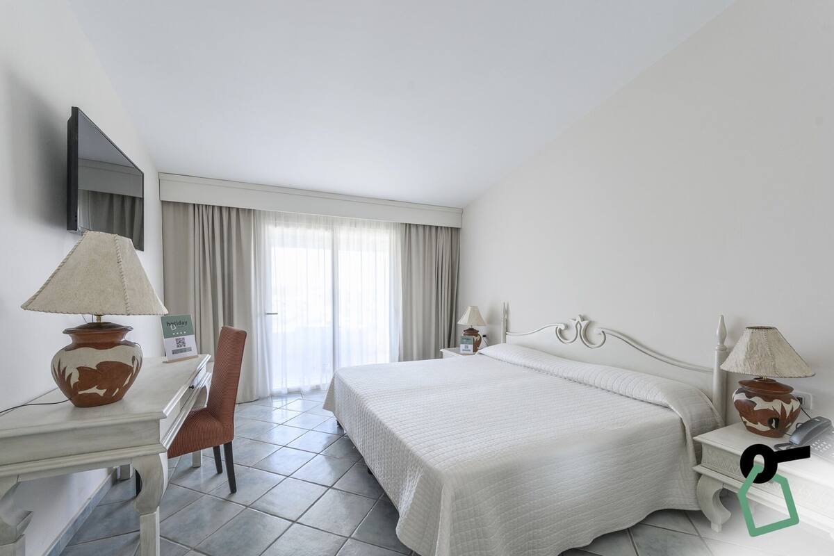 A hotel room with a bed, two bedside tables, a chair, a desk, a lamp, a mirror, and a window with curtains.