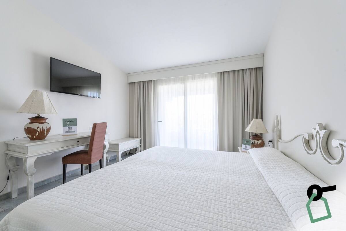 A hotel room with a large bed, a desk with a chair, a TV mounted on the wall, and a window with curtains.