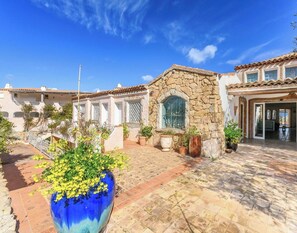 A courtyard with a stone wall, a large blue vase with yellow flowers, and a building with a tiled roof. - Hotiday Room Collection - Porto Cervo (Arzachena)