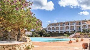 Zona de piscina con pared de piedra y plantas en macetas, un edificio grande con varios balcones y cielo despejado.