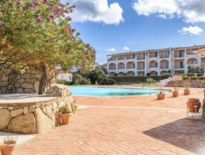A swimming pool area with a stone wall and potted plants, a large building with multiple balconies, and a clear sky. - Hotiday Room Collection - Porto Cervo (Arzachena)
