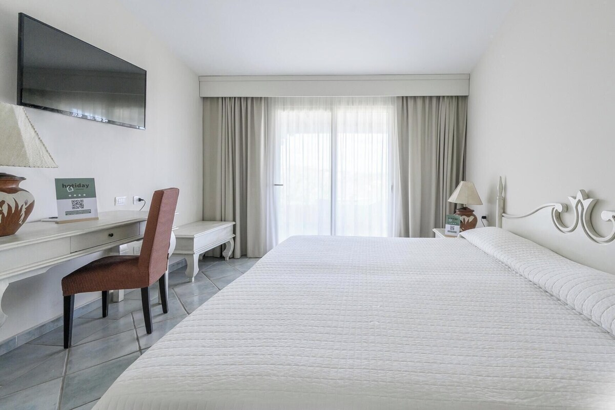 A hotel room with a large bed, a desk with a chair, a flat-screen TV, and a window with curtains.