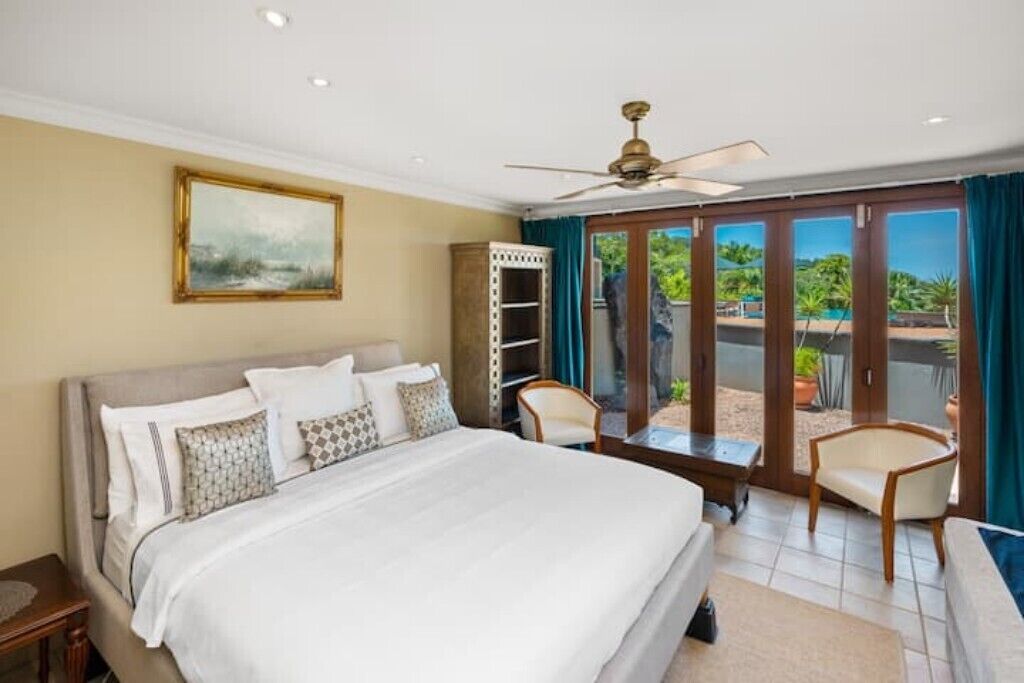 Une chambre dotée d’un grand lit, d’un ventilateur de plafond, d’une armoire et offrant une vue sur une piscine et un espace verdoyant, le tout visible à travers une porte-fenêtre coulissante.
