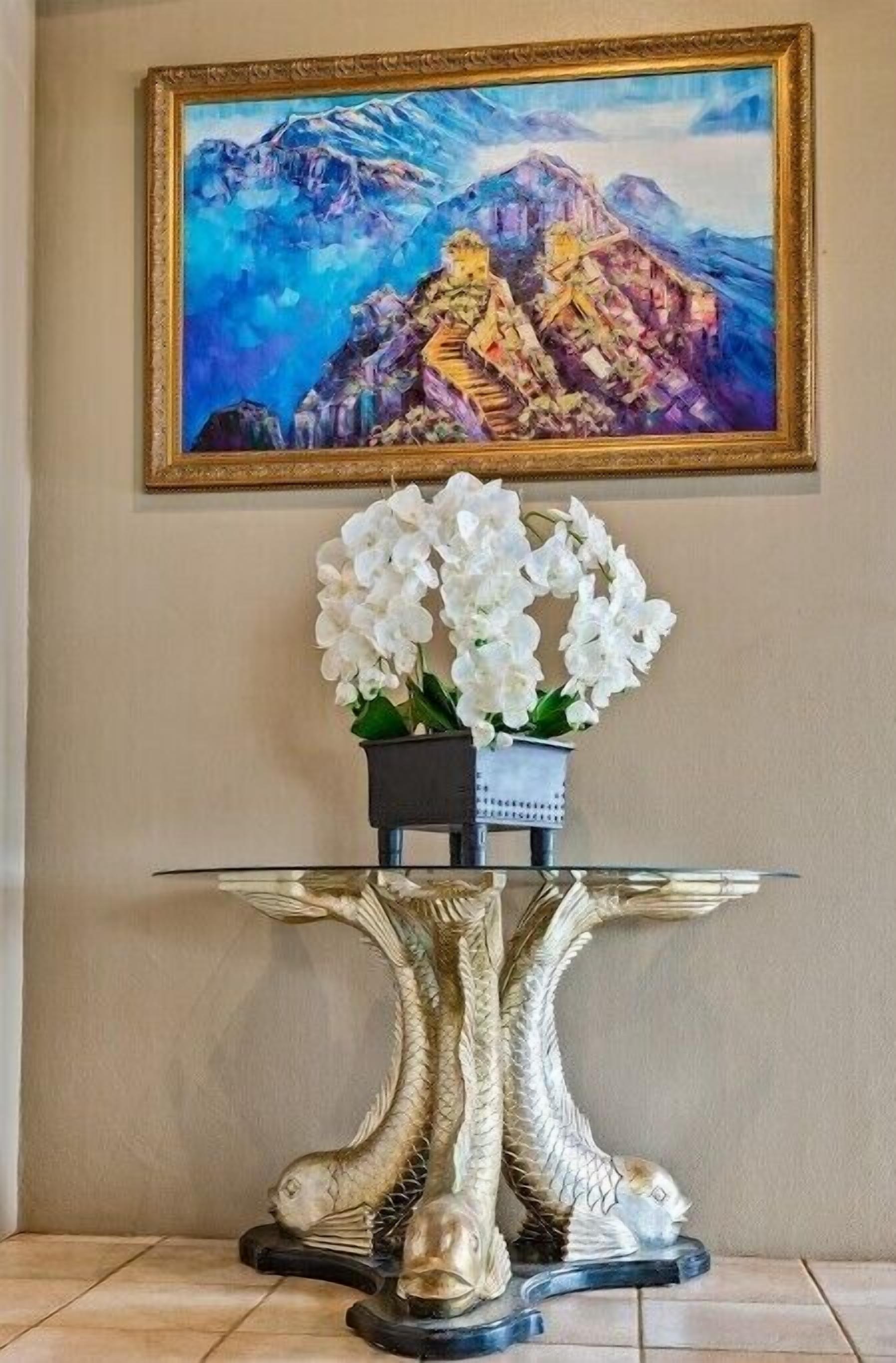 Une table dont le piétement est orné d’une sculpture de poisson, un vase rempli d’orchidées blanches et un tableau encadré placé au-dessus.