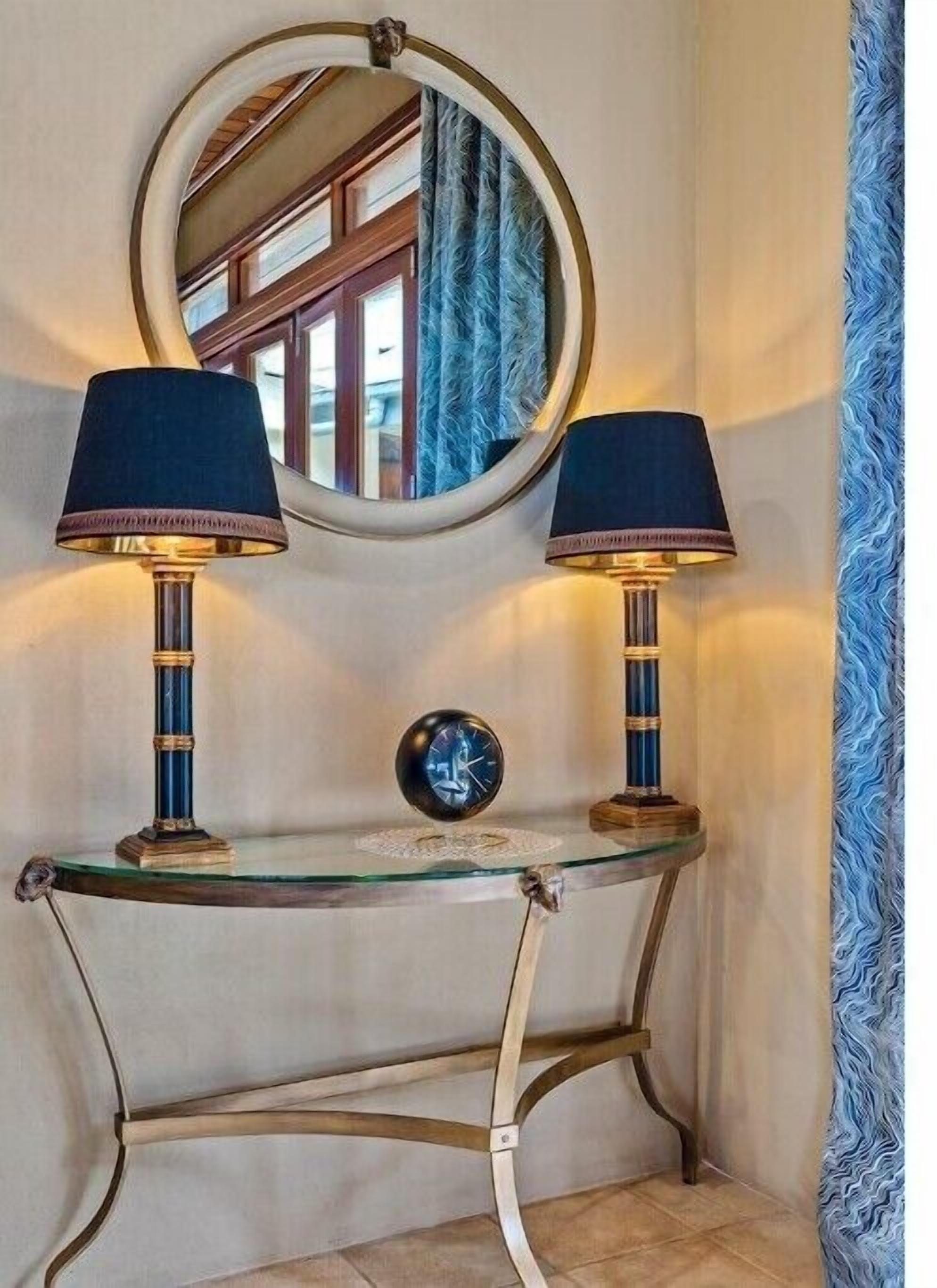 A corner with a round mirror, two lamps, and a small clock on a glass table.
