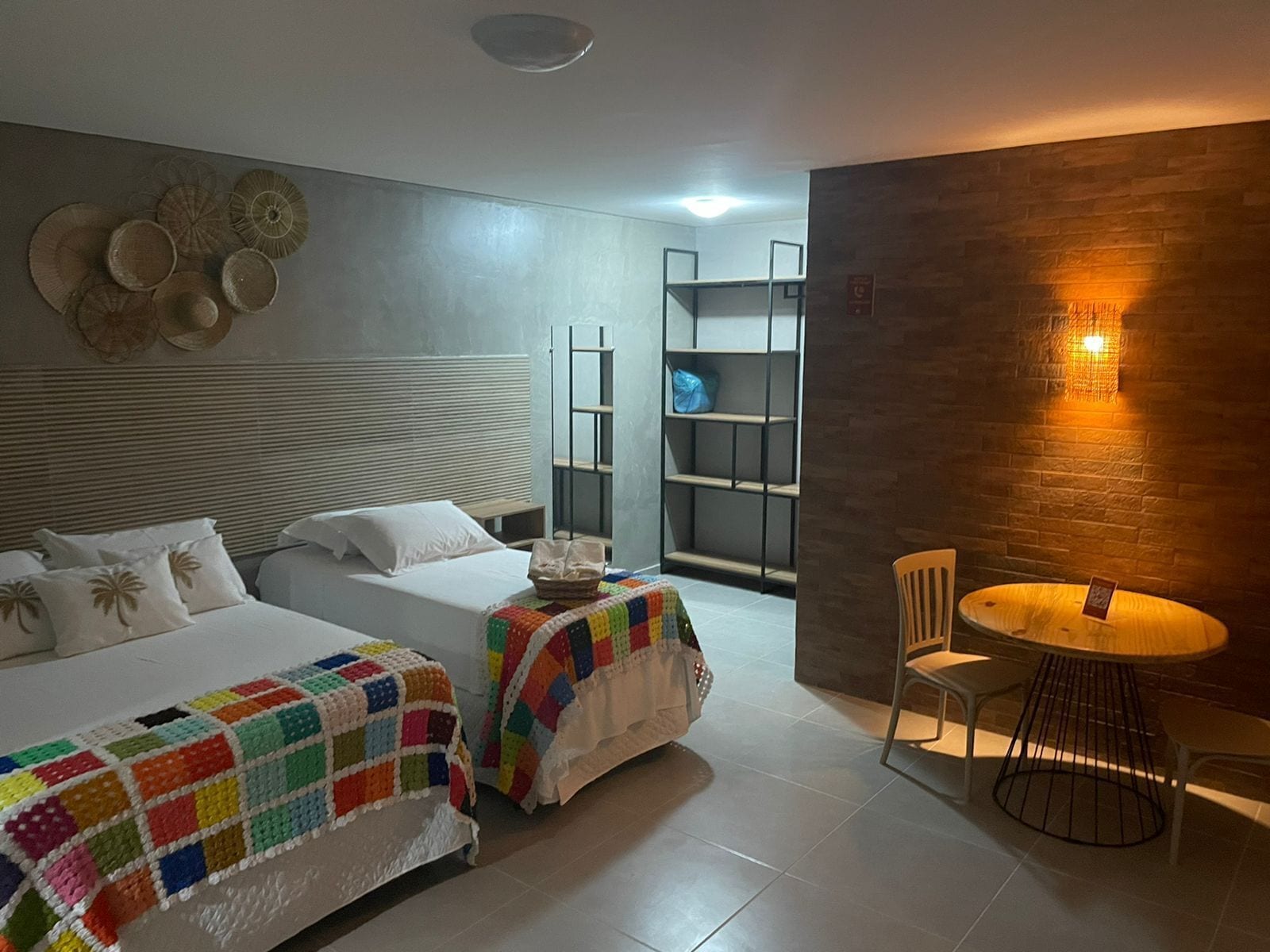 Une chambre d’hôtel avec deux lits, une petite table et une chaise. Il y a une étagère dans un coin et un luminaire fixé au mur.