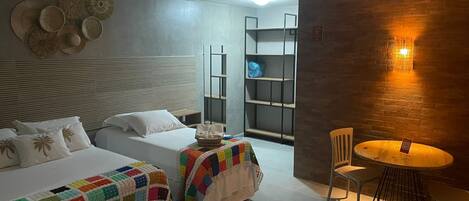 Une chambre d’hôtel avec deux lits, une petite table et une chaise. Il y a une étagère dans un coin et un luminaire fixé au mur.