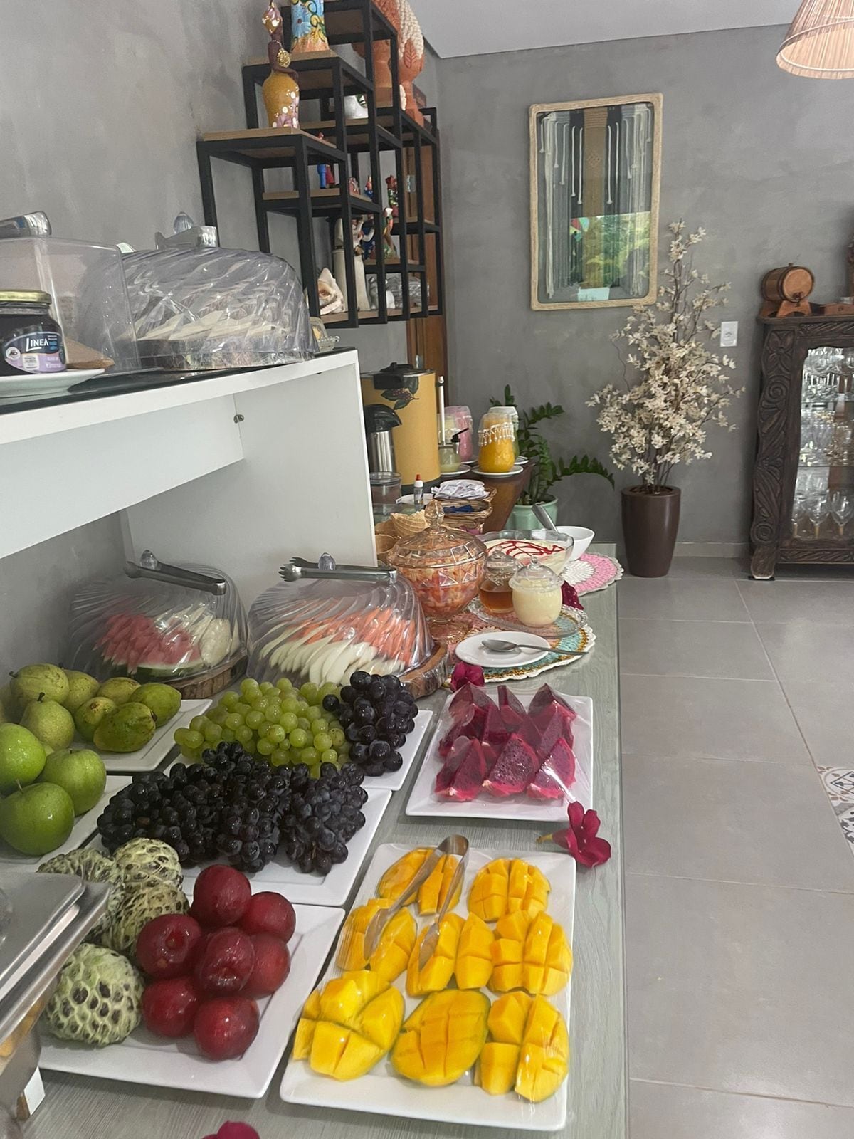 A fruit and snack display with various fruits like apples, grapes, and mangoes, and a shelf with decorative items and a mirror.