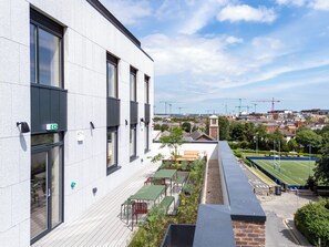 A modern building with large windows, a rooftop garden with seating, and a view of a sports field and construction cranes in the distance.