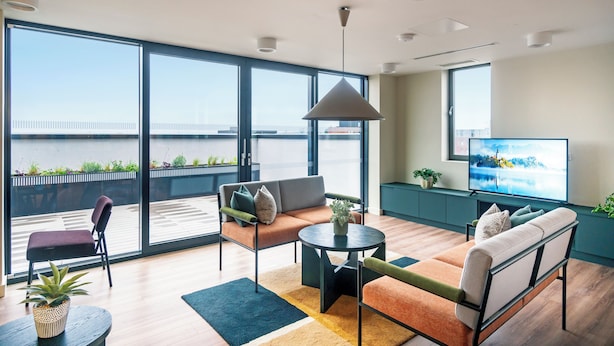 A modern living room with a sofa, armchairs, a coffee table, and a TV. Large windows offer a view of the city. - Niche Living Rathmines (Dublin)