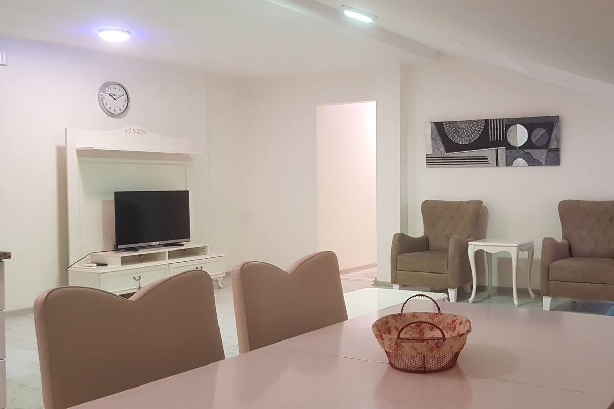 A modern living room with a dining area, a flat-screen TV, two armchairs, a sofa, and a wall clock.