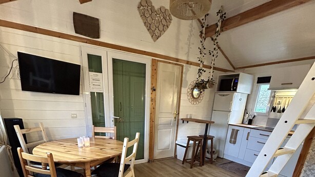 Un intérieur chaleureux et rustique avec une table à manger en bois, des chaises, un petit coin cuisine et une mezzanine accessible par une échelle.