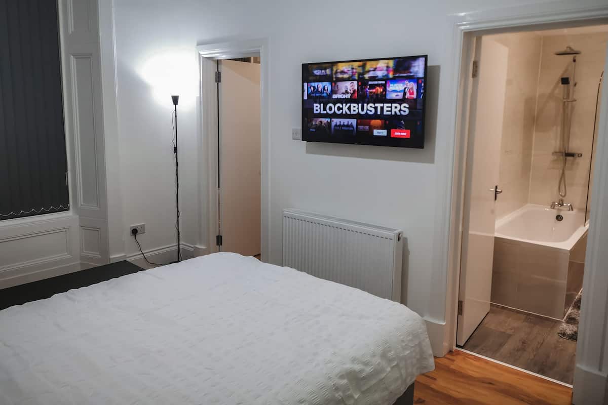 A hotel room with a bed, a TV displaying 'BLOCKBUSTERS', a radiator, a shower, and a door leading to another room.