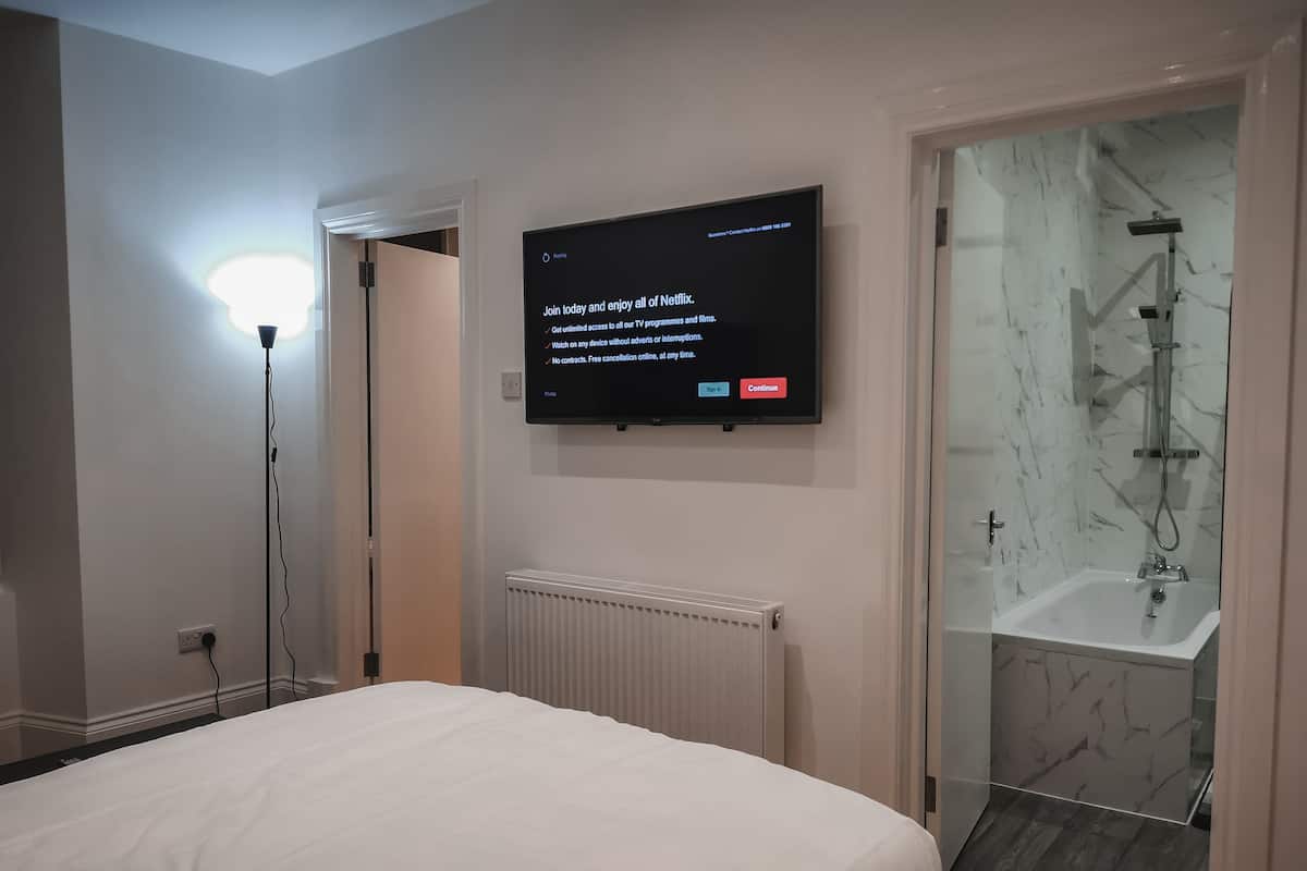A modern hotel room with a large bed, a mounted TV displaying a Netflix sign, a radiator, and a glass-enclosed shower area.