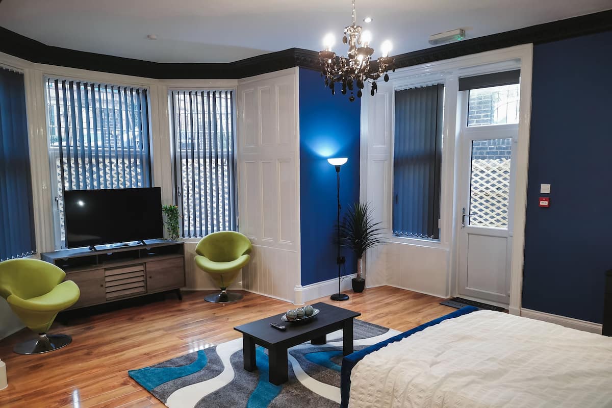 A modern living room with a flat-screen TV, two green chairs, a coffee table, and a large bed.