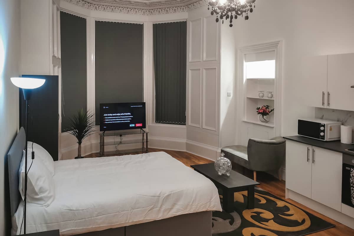 A modern hotel room with a large bed, a desk, a TV, a kitchenette, and a view of the city.
