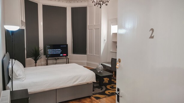 A bedroom with a bed, a desk, a TV, a chandelier, and a door with a number. - SH Apartments - Dixon Street (Glasgow)