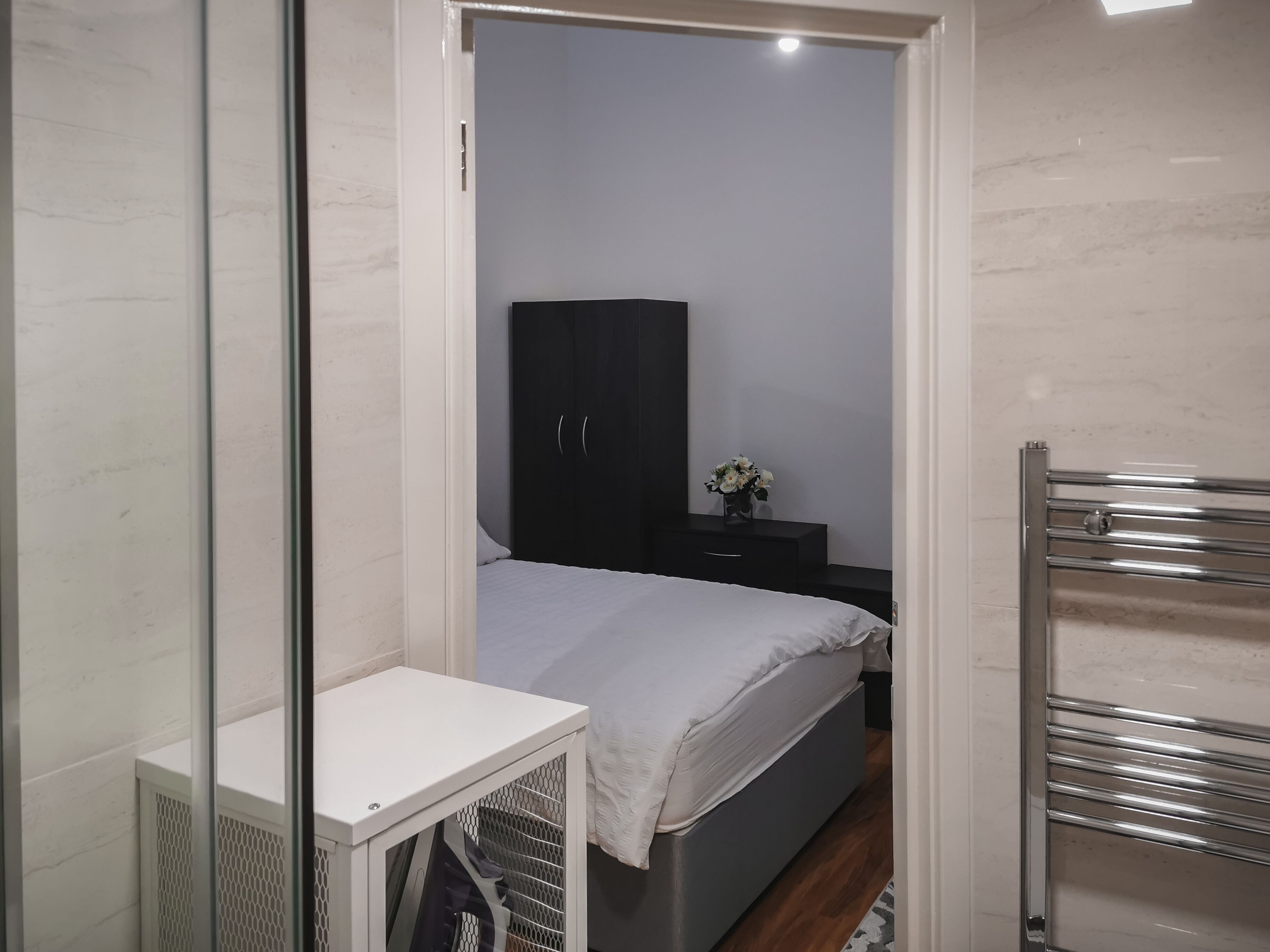 Une petite chambre à coucher moderne, équipée d’un lit, d’une armoire, d’une table de chevet agrémentée d’un vase de fleurs et d’un radiateur encastré.