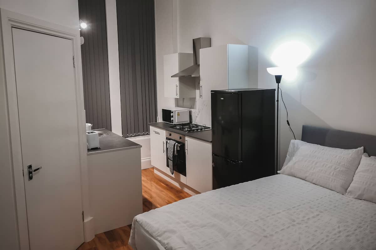 A compact bedroom with a bed, a small kitchenette with a refrigerator, microwave, and stove, and a door leading to a bathroom.