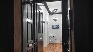 A hallway with a black door, a black mat at the entrance, a washing machine, and a television mounted on the wall. - SH Apartments - Dixon Street (Glasgow)