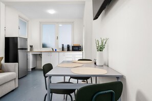 A modern kitchen with white cabinets, a black refrigerator, and a microwave. There is a dining table with chairs and a potted plant on the windowsill. - Otium Rentals Comfort Studio in Condado (San Juan)