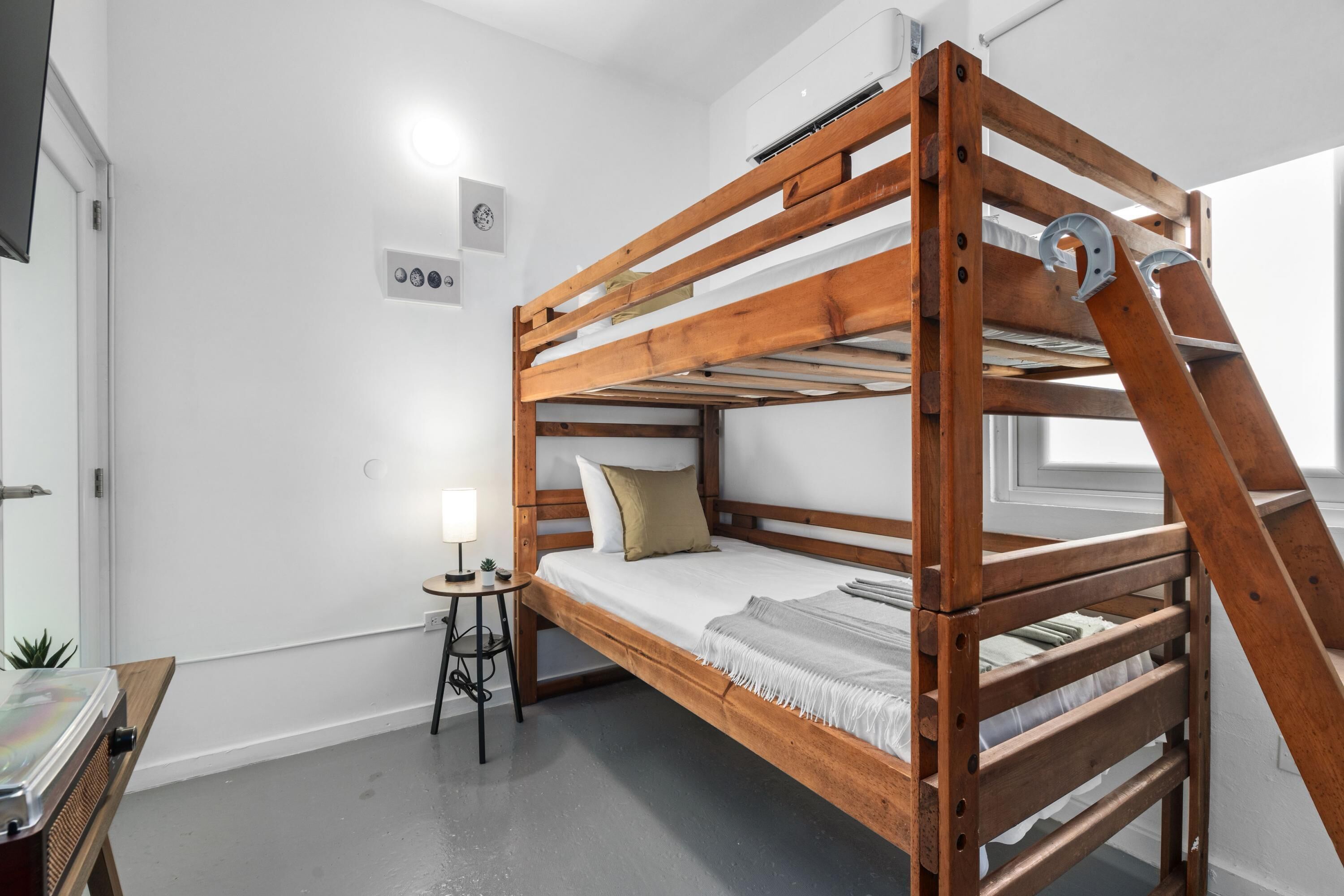 A bunk bed with a wooden frame, a small bedside table with a lamp, and a window in the background.