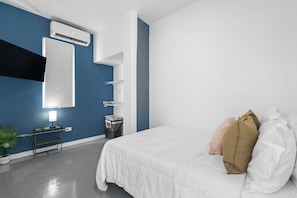 A modern hotel room with a large bed, a bedside table with a lamp, a wall-mounted air conditioner, and a small plant. - Otium Rentals Perfect Family Getaway Place (San Juan)