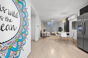 A modern kitchen with a stainless steel refrigerator, a dining area with a white table and chairs, and a living room with a sofa and a decorative wall.