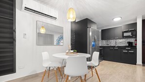 Egy modern konyha, étkezővel, melyben egy kerek asztal fehér székekkel, beépített tűzhely és sötét színű konyhaszekrények találhatók.