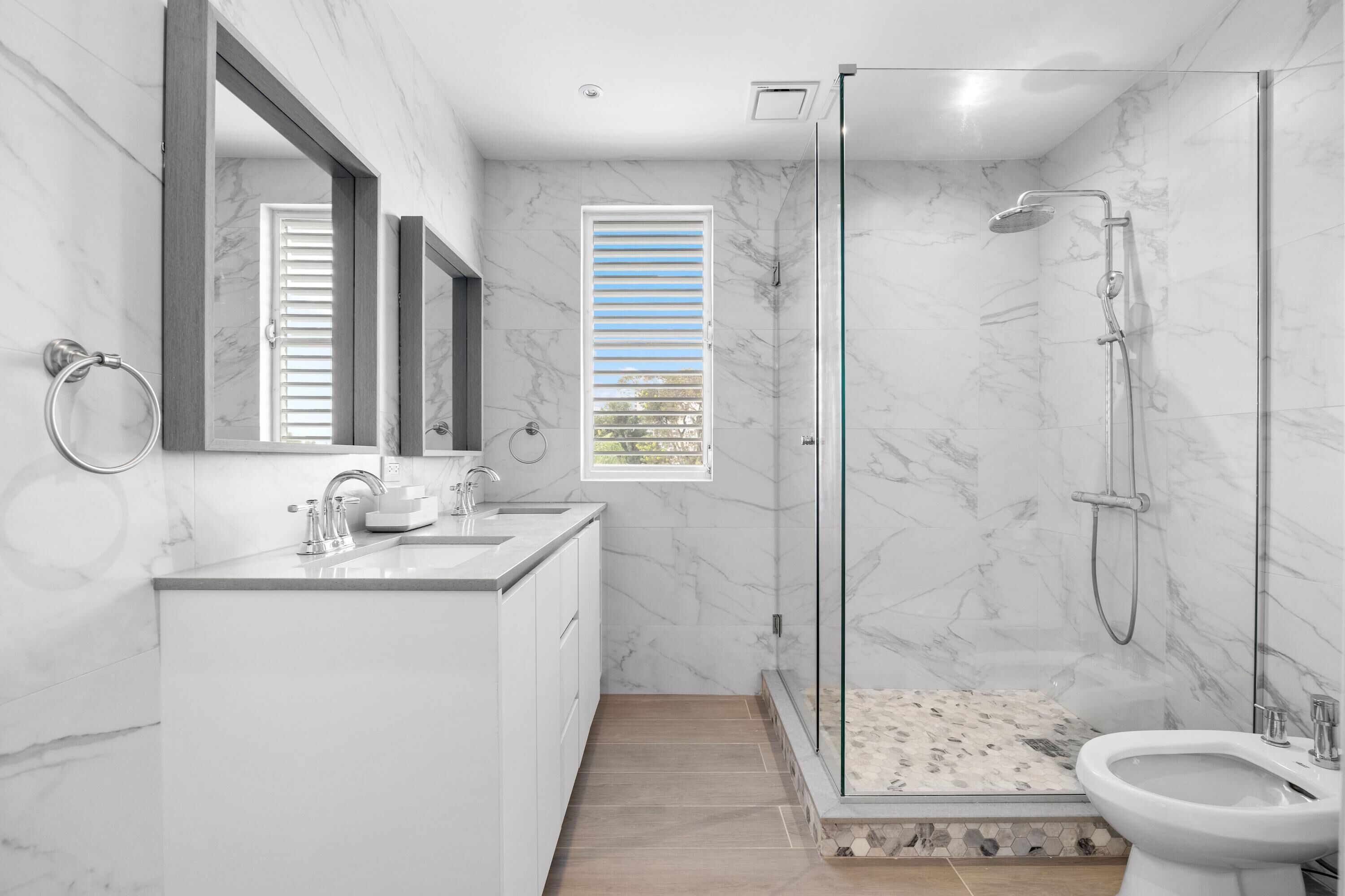 Un baño moderno con ducha envuelta en vidrio, lavamanos con argolla para toallas y ventana con persianas.