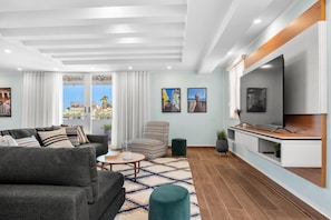 A modern living room with a sofa, armchair, and coffee table, a TV on a wooden stand, and a view through large windows. - Otium Rentals Casa Elena Rental for Families (San Juan)